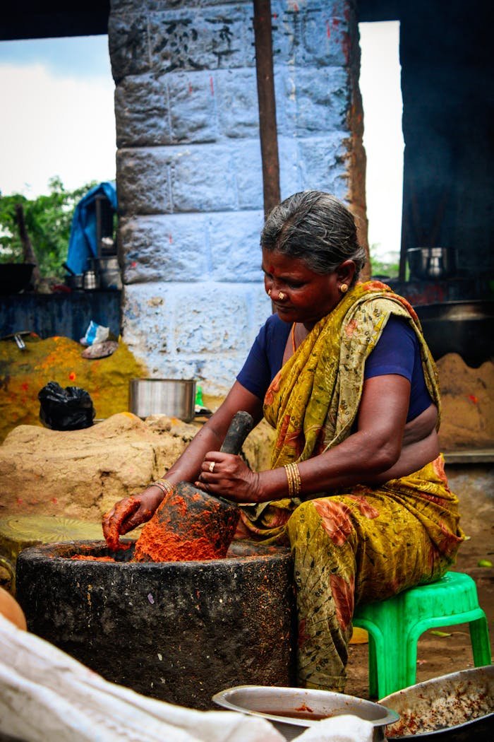 Full body senior ethnic female in colorful traditional clothes sitting on plastic stool in messy courtyard and powdering spices in big stone bowl